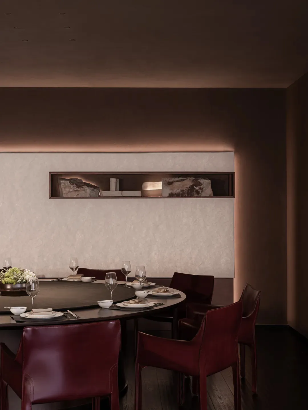 Dining room with a round table set with wine glasses and a vase, surrounded by chairs in a brown color scheme, with dim lighting creating a cozy atmosphere