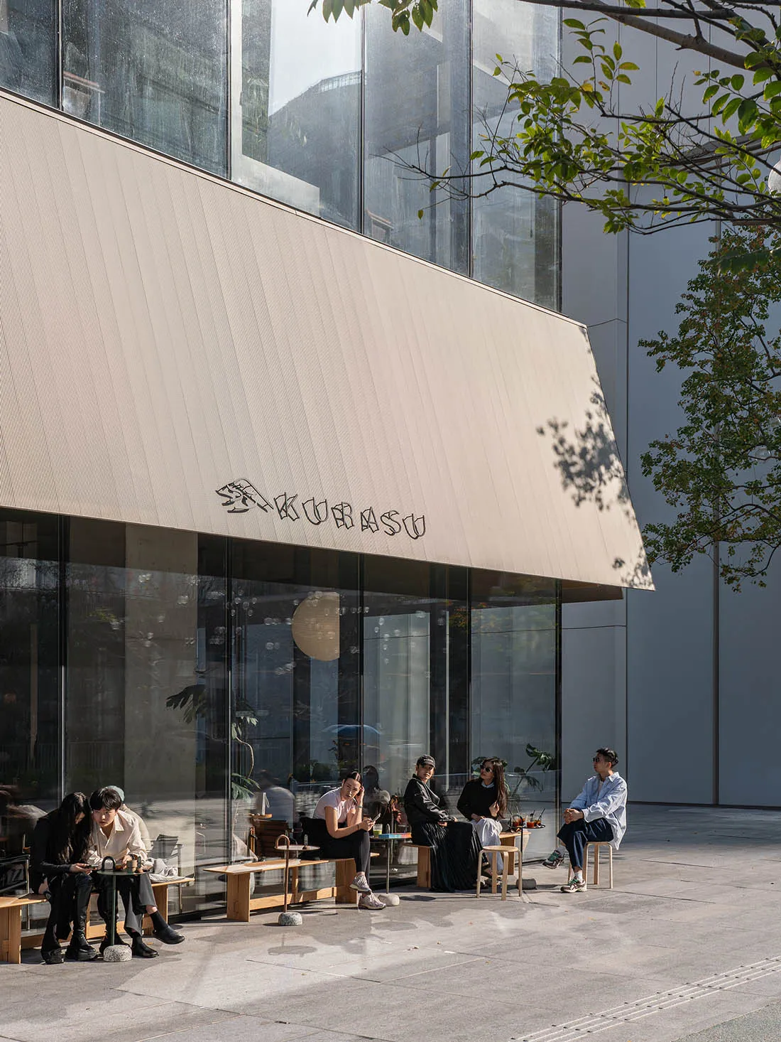 A sunny outdoor cafe scene with a white-roofed building, large window, and people sitting on chairs and benches