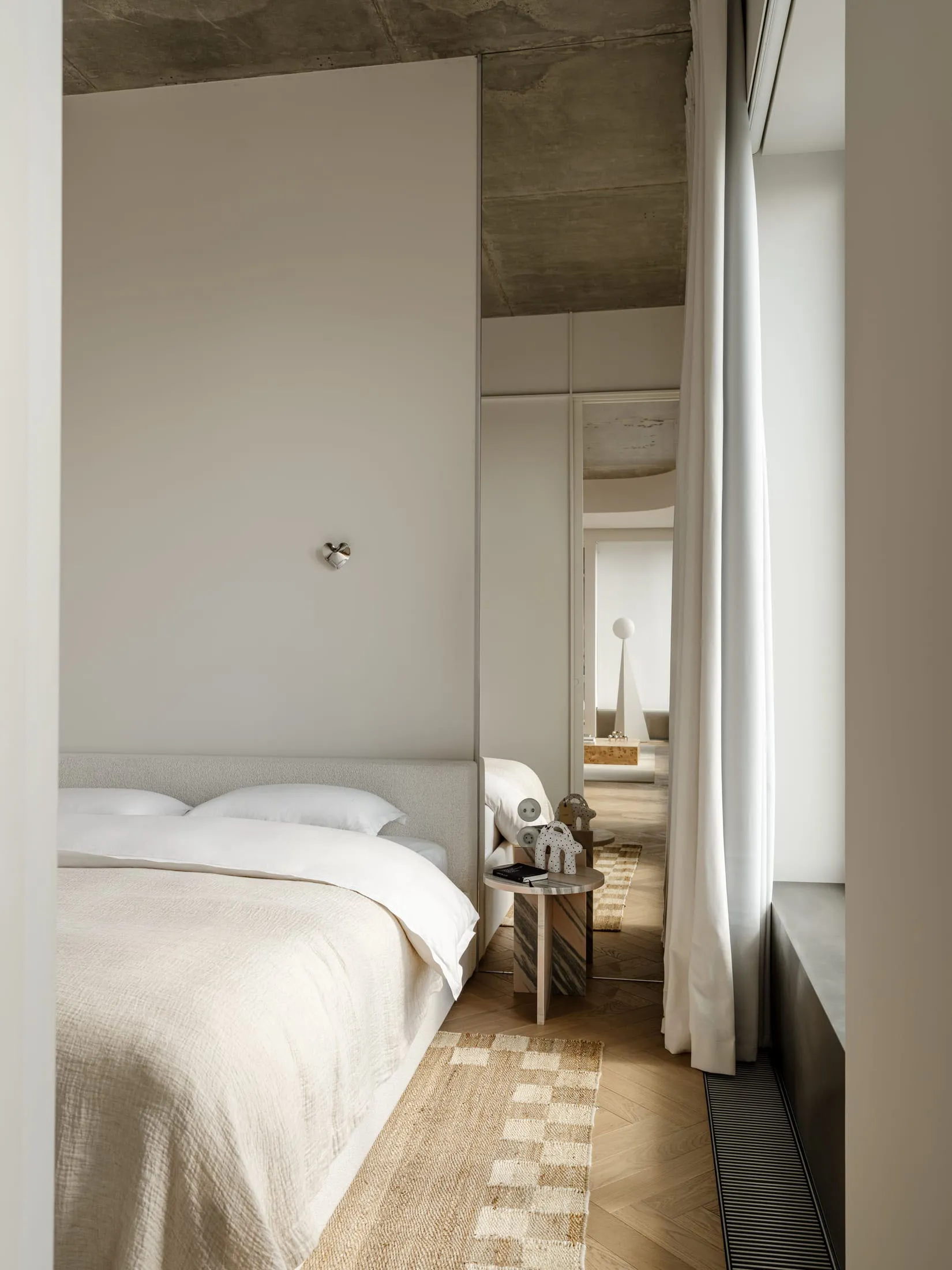A well-lit bedroom with a bed, a mirror, and a table, surrounded by white walls and wood accents