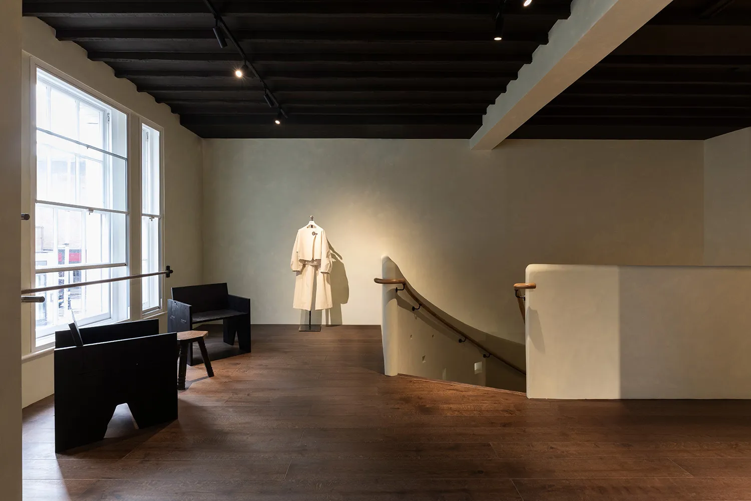 A modern lobby with a wooden floor, white walls, a staircase, and seating areas with chairs and a bench, lit by dim lighting