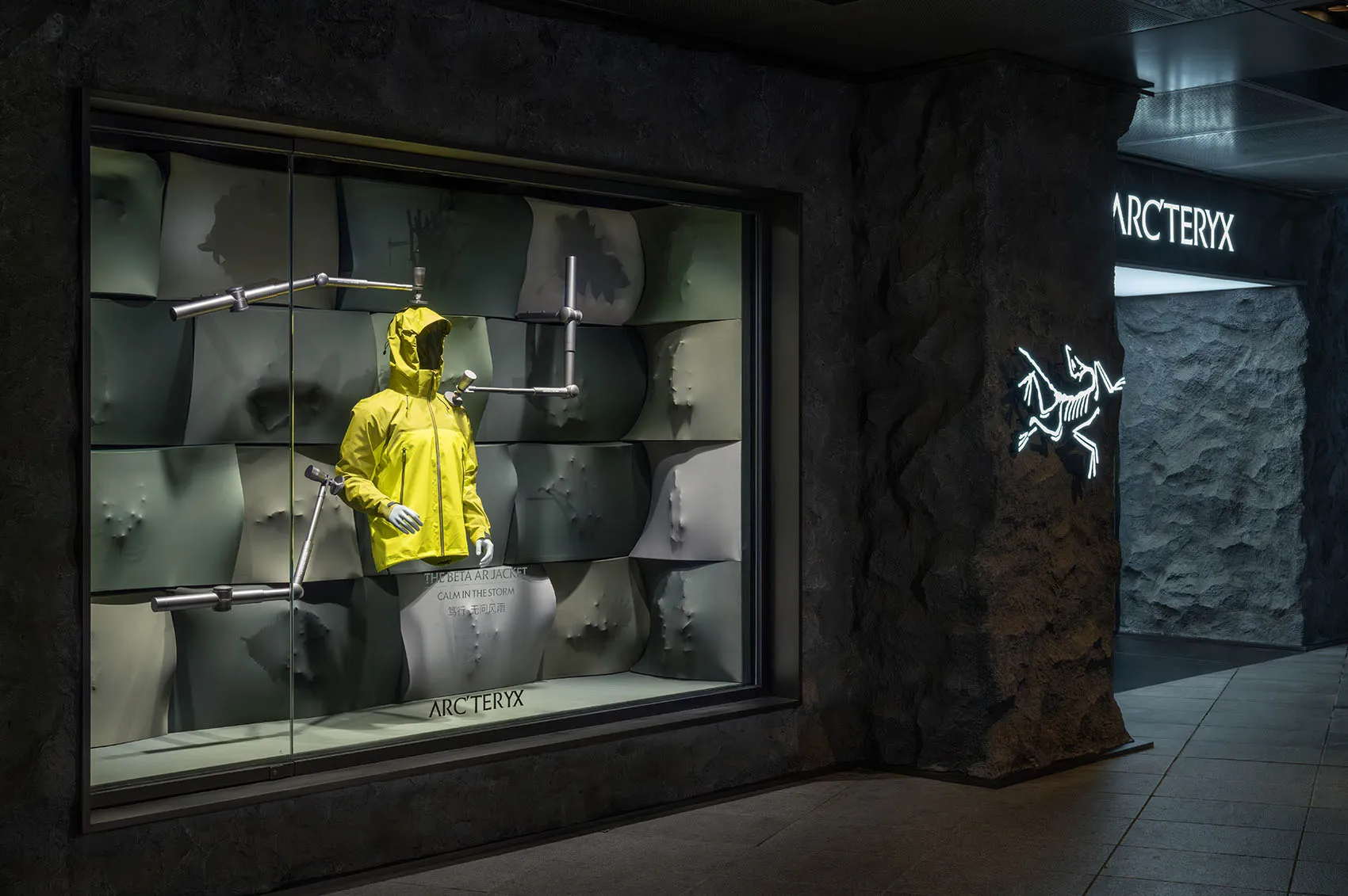 A mannequin wearing a yellow jacket stands in front of a stone wall in a store display, creating a striking contrast