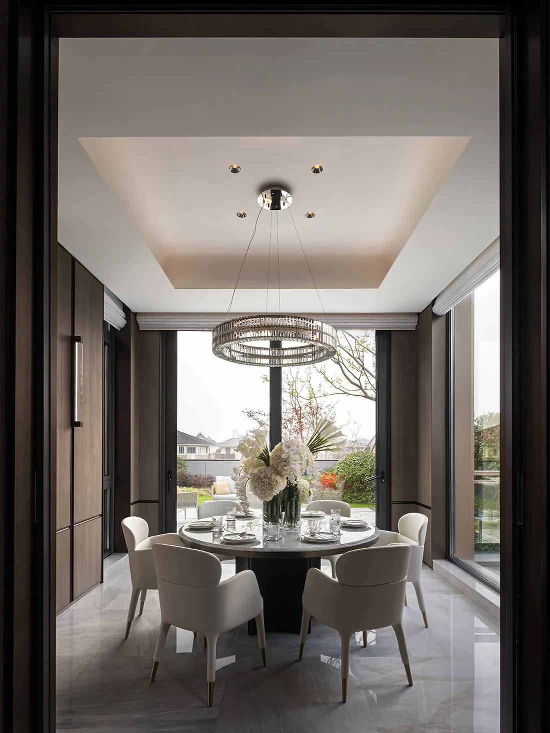 Dining room with a large wooden table and chairs, set with plates, wine glasses, and a vase of flowers, under a chandelier in a well-lit space