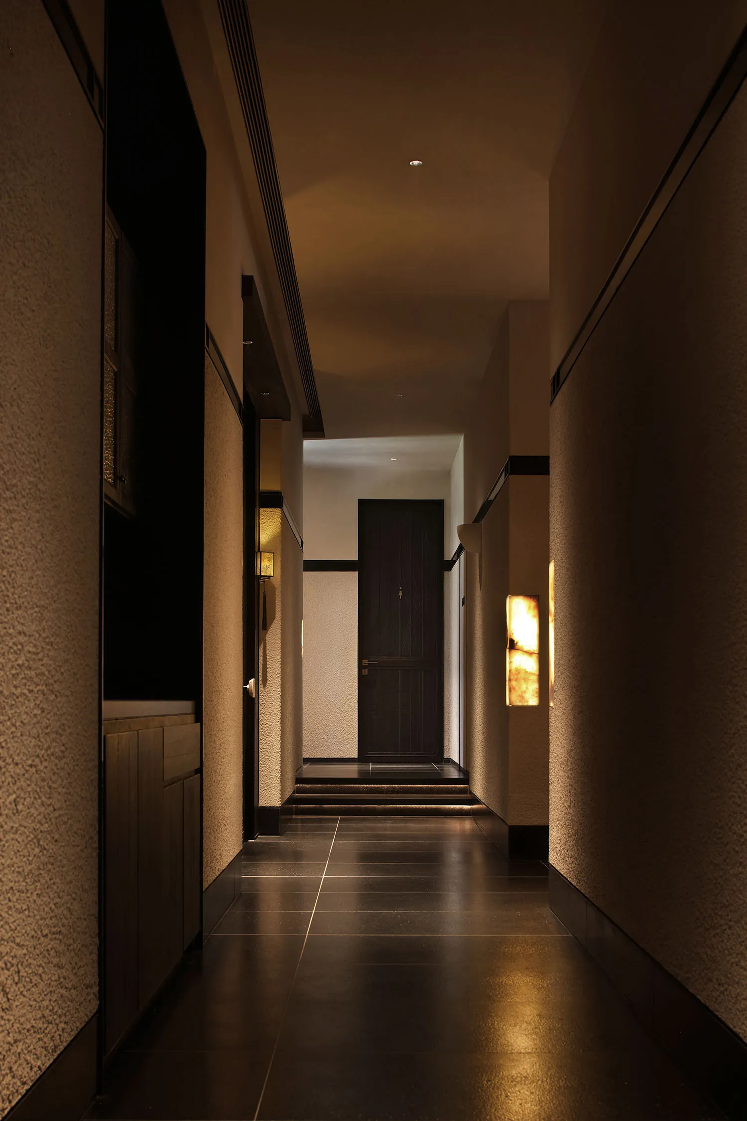A modern hallway with a doorway, staircase, and concrete walls, lit by a wall light