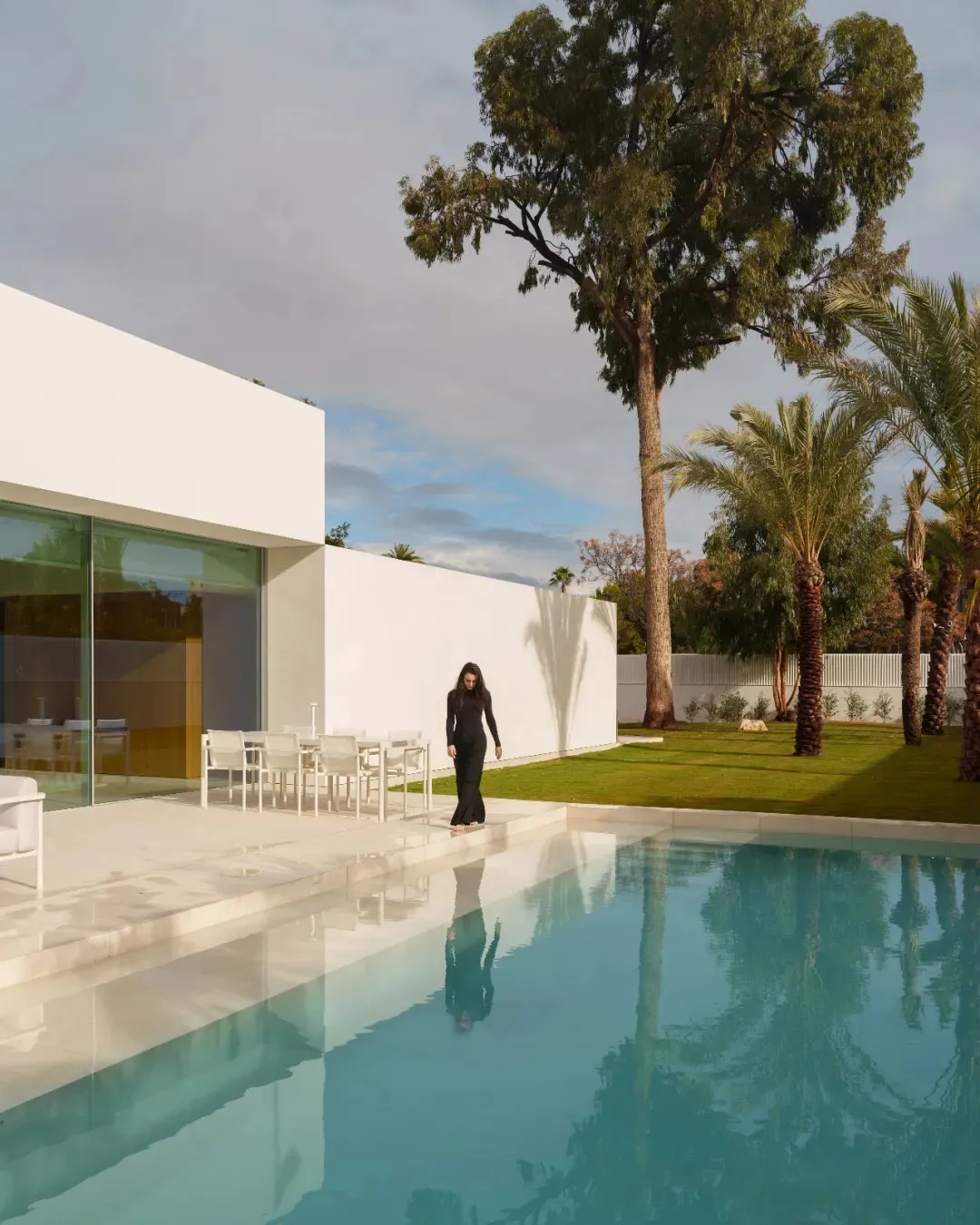A modern outdoor living space with a pool surrounded by a white wall, a patio with a dining table and chairs, and potted plants, with a person standing near the pool, lit by natural sunlight