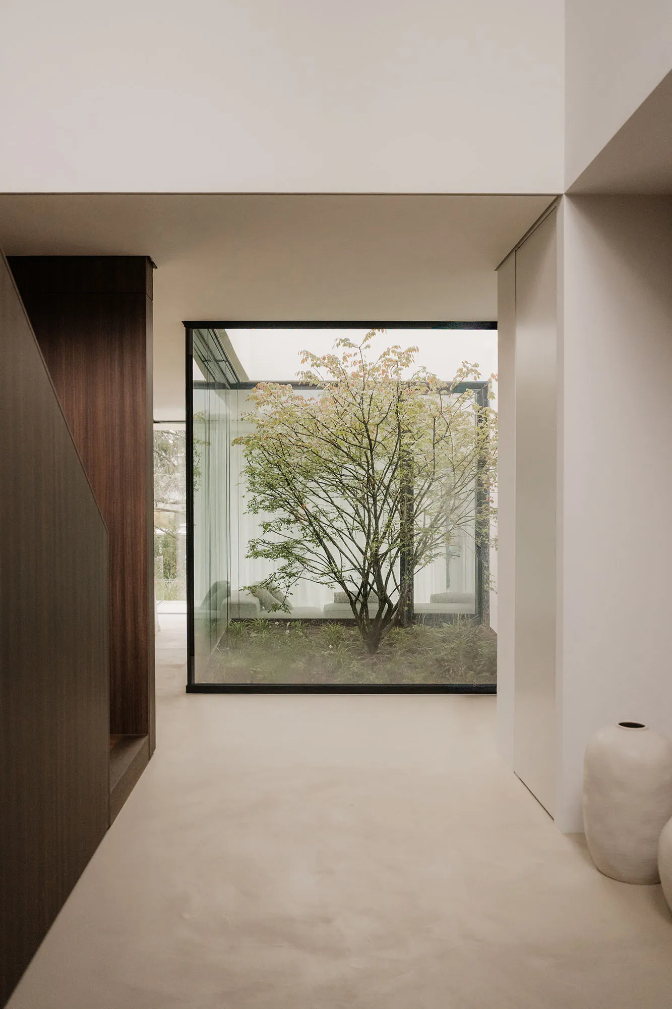 A living room with a couch, chair, and wooden floor, featuring a large glass window with a tree outside, allowing natural light to enter and creating a bright and welcoming atmosphere