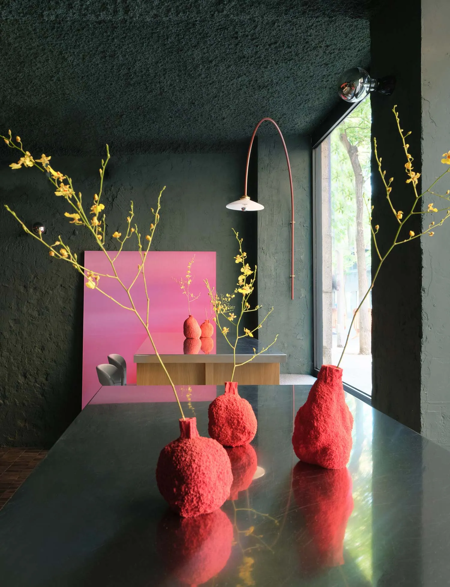 A modern living area with a dining table, red vases, and flowers, surrounded by chairs and a potted plant, with a warm and inviting ambiance