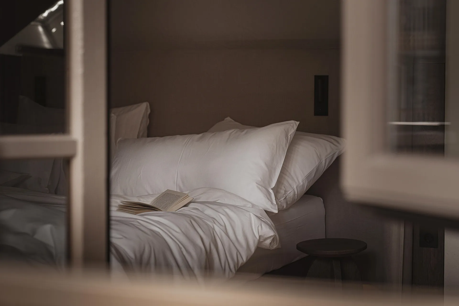 Cozy bedroom with a bed, chair, and book, lit by natural light from the window, with wood and fabric visible