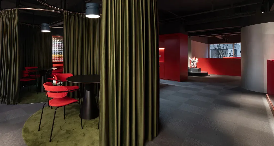 Modern restaurant interior featuring olive green velvet curtains, bright red upholstered chairs, black round tables, and a checkered carpet floor with dramatic red accent walls.