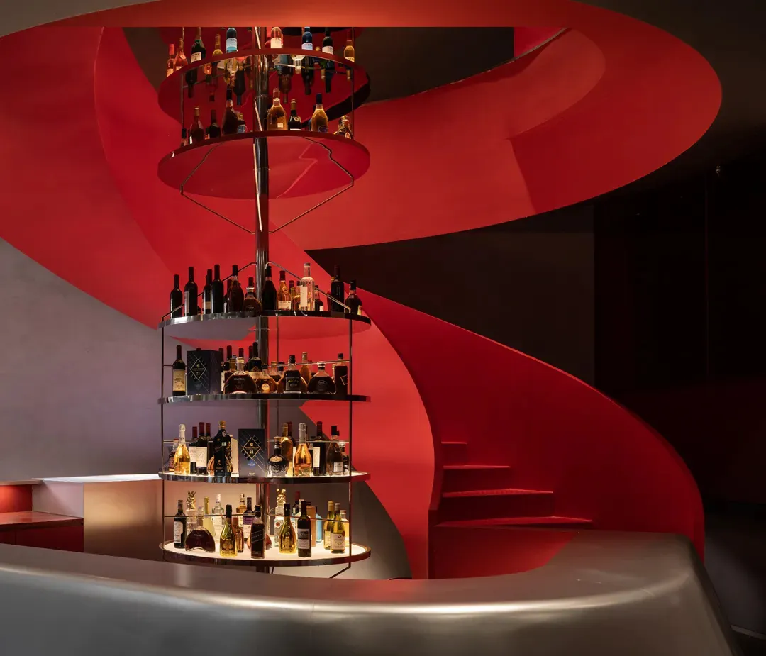 A dimly lit bar with a red metal spiral staircase and a shelf filled with liquor bottles, creating a sophisticated and inviting atmosphere