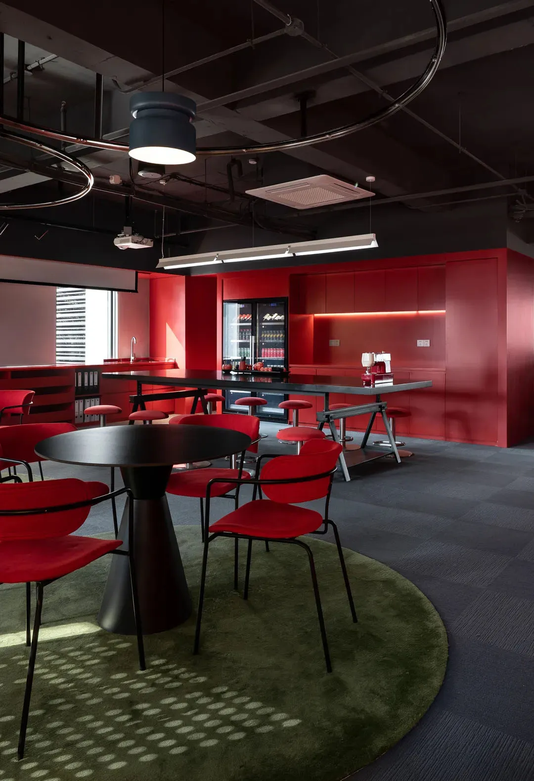 A dimly lit dining kitchen with a red carpet, a red table, and a modern kitchen area with a sink and refrigerator, surrounded by chairs