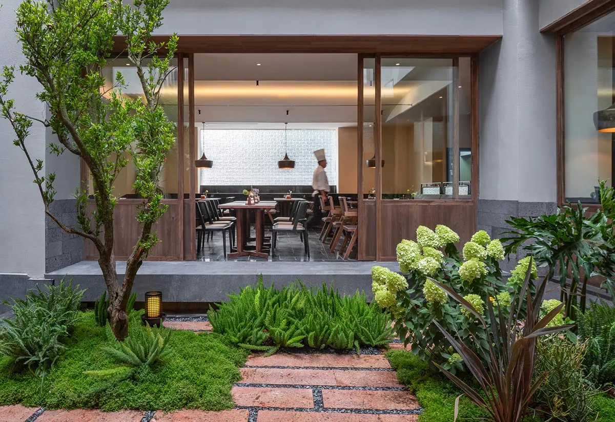 A modern restaurant with a dining area featuring a wooden table and chairs, and a kitchen with metal appliances and a neutral color scheme