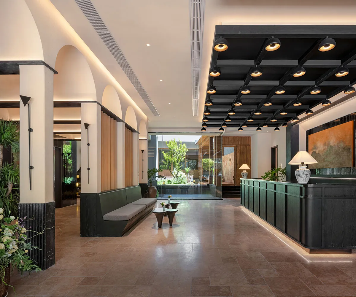 A modern hotel lobby with a large couch, bench, and dining table, surrounded by potted plants and illuminated by natural light