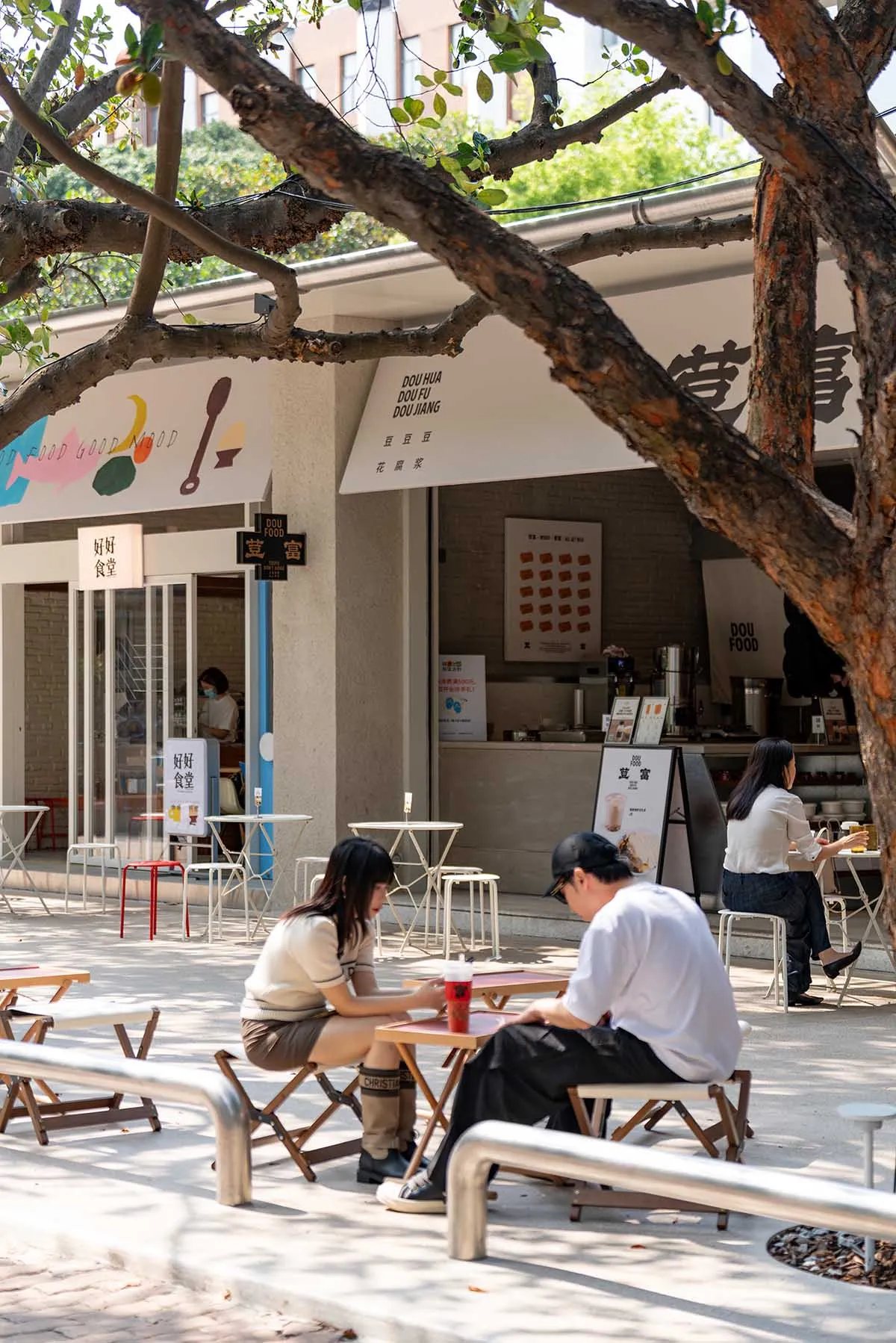 A casual outdoor dining area with people sitting at tables and chairs under a tree, surrounded by a modern restaurant with wood and metal materials, lit by natural sunlight