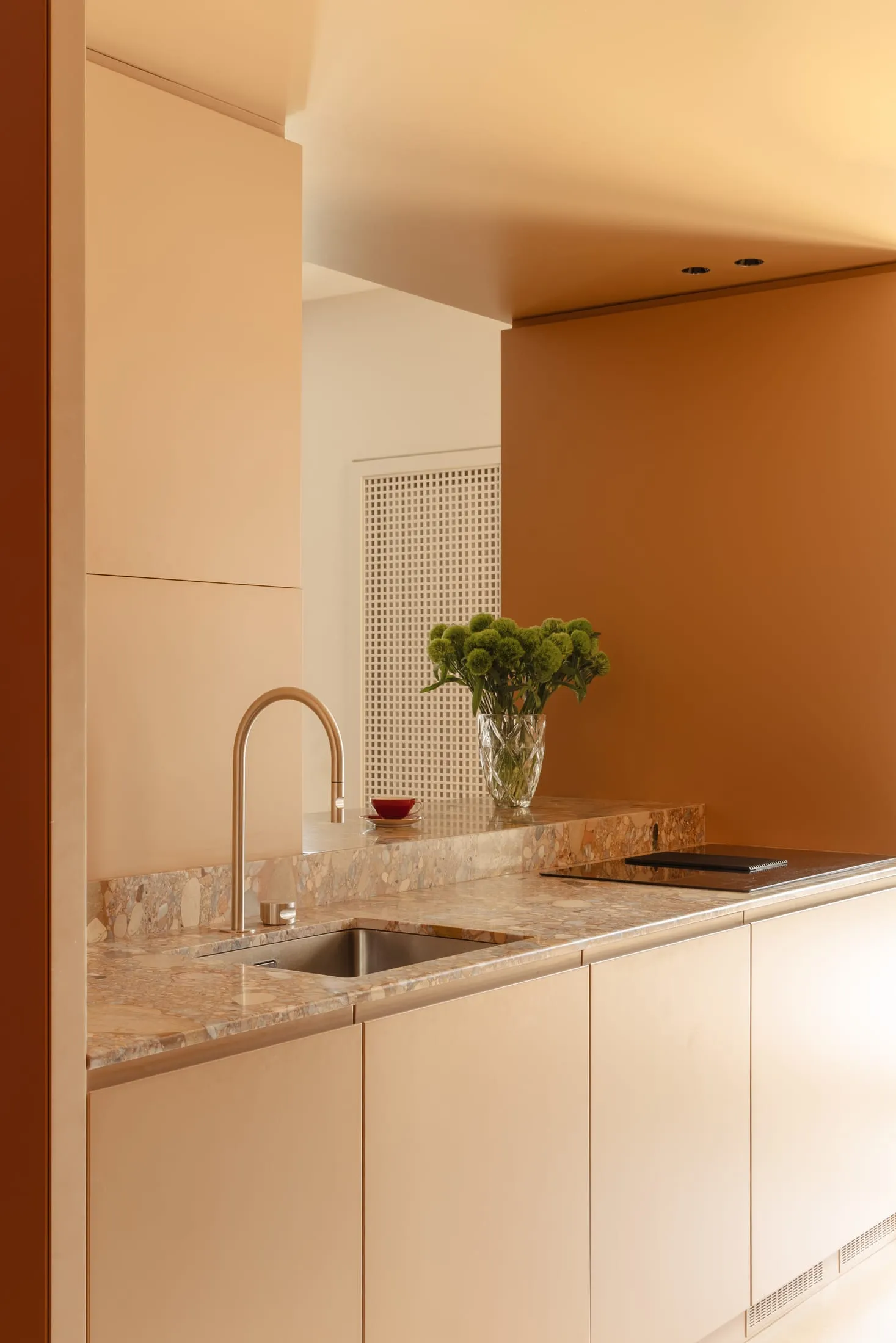 Modern kitchen with a marble countertop, a brown wall, a metal sink, a glass vase, and a potted plant, with soft and warm lighting