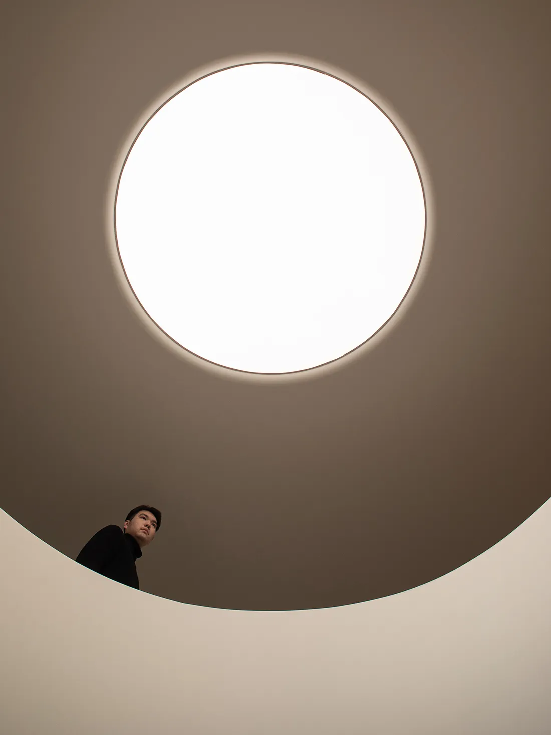 A man stands under a large round white light fixture in a modern minimalist room with a circular design and clean aesthetic