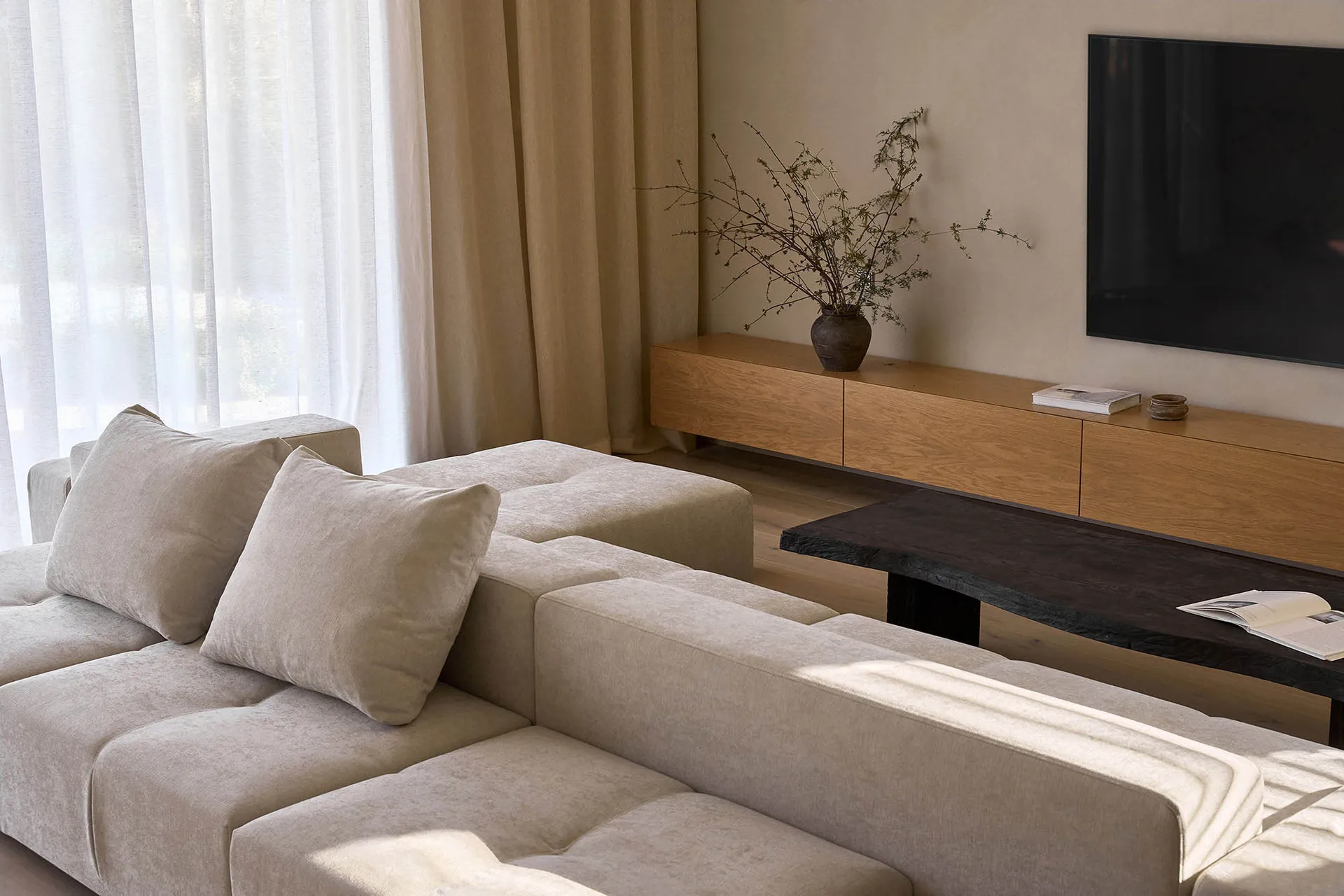 A modern living room with a large TV, white couch and chair, a potted plant, and natural light from the windows