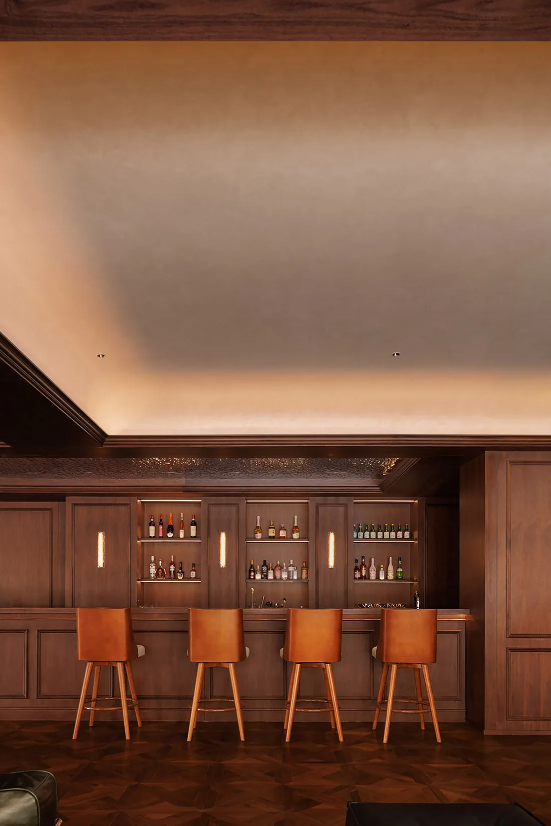 Cozy bar area with a wooden counter, brown chairs, and dim lighting, surrounded by bottles and seating for patrons