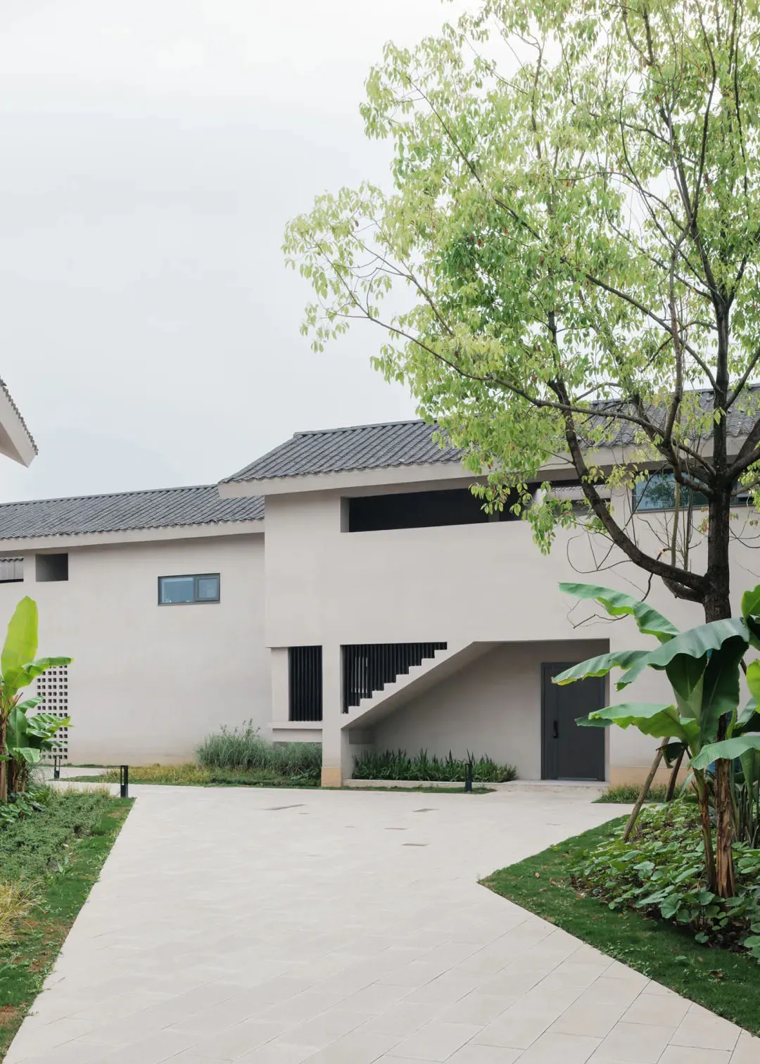 A modern house with a white exterior, large window, and staircase, set in a residential area with a tree in the background, under natural sunlight