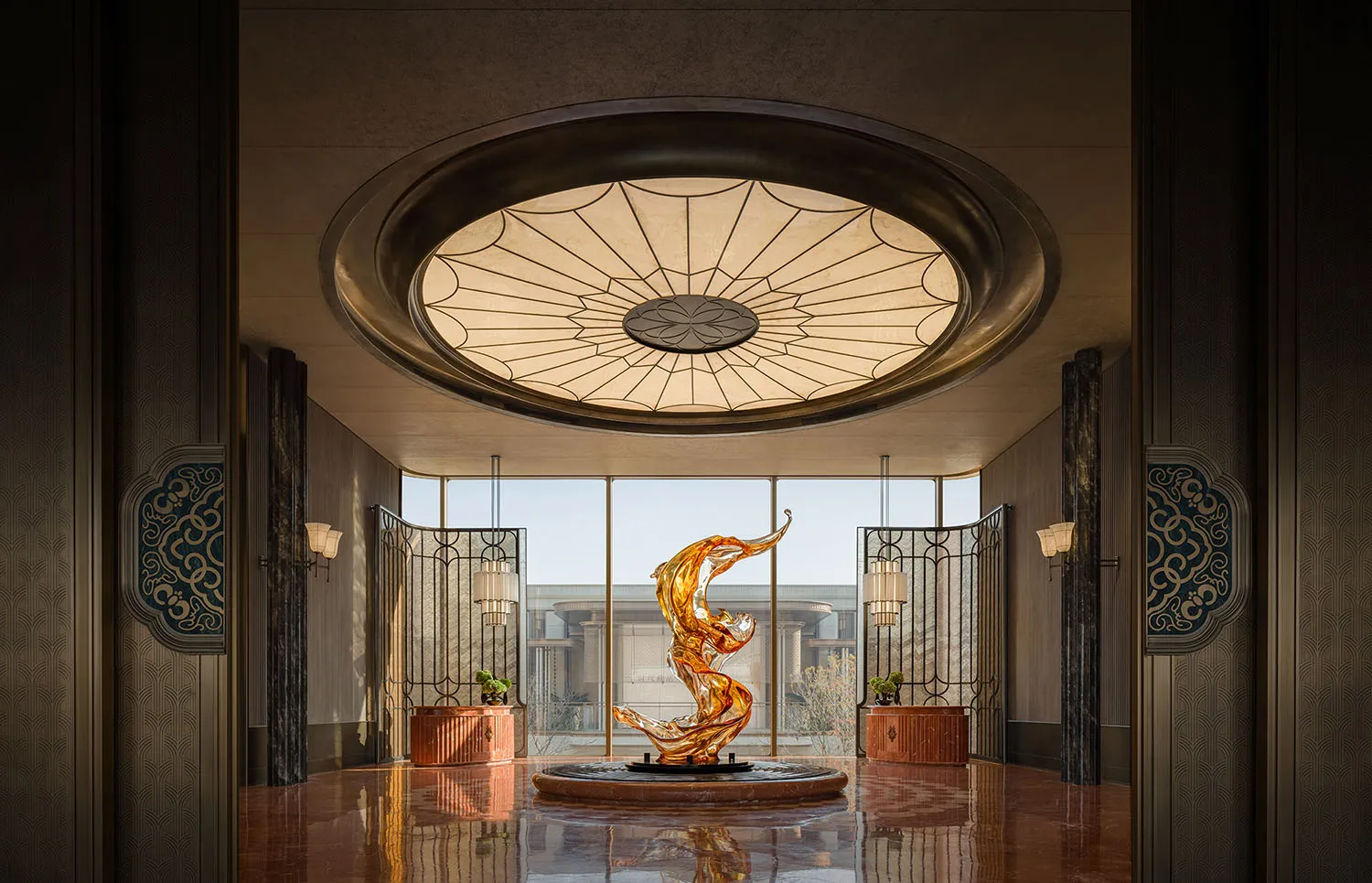 Bright and inviting lobby with a glass ceiling, a circular light fixture, a large sculpture, and a fountain, illuminated by natural light