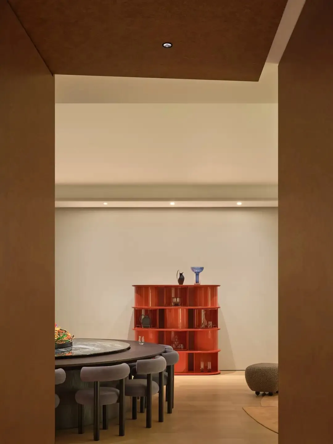 A modern study with a wooden floor, a dining table, a red bookshelf, and a chair, lit by natural sunlight