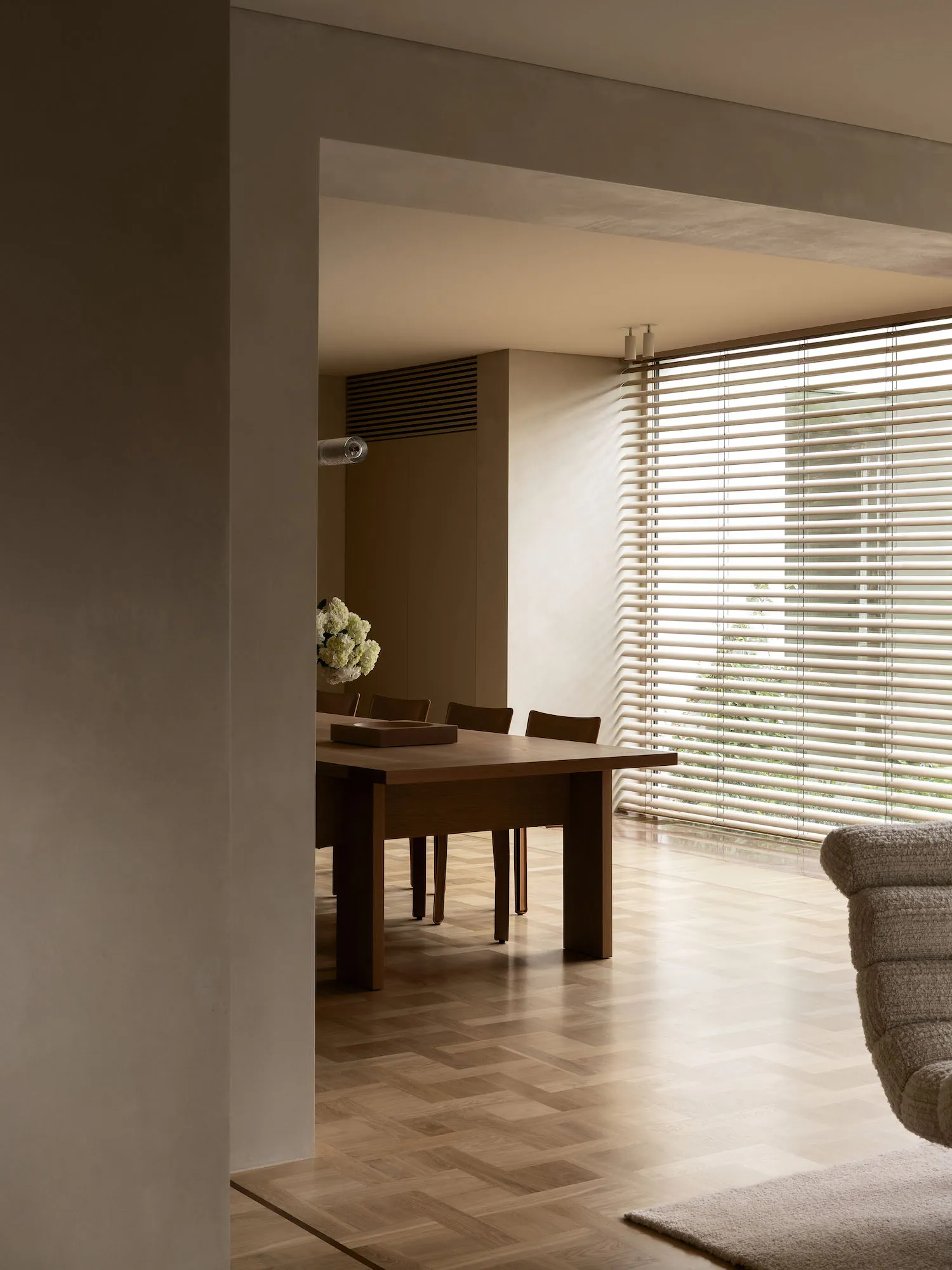 A well-lit living room with a wooden floor, a dining table, chairs, a couch, and a vase, creating a cozy atmosphere with natural light from the windows