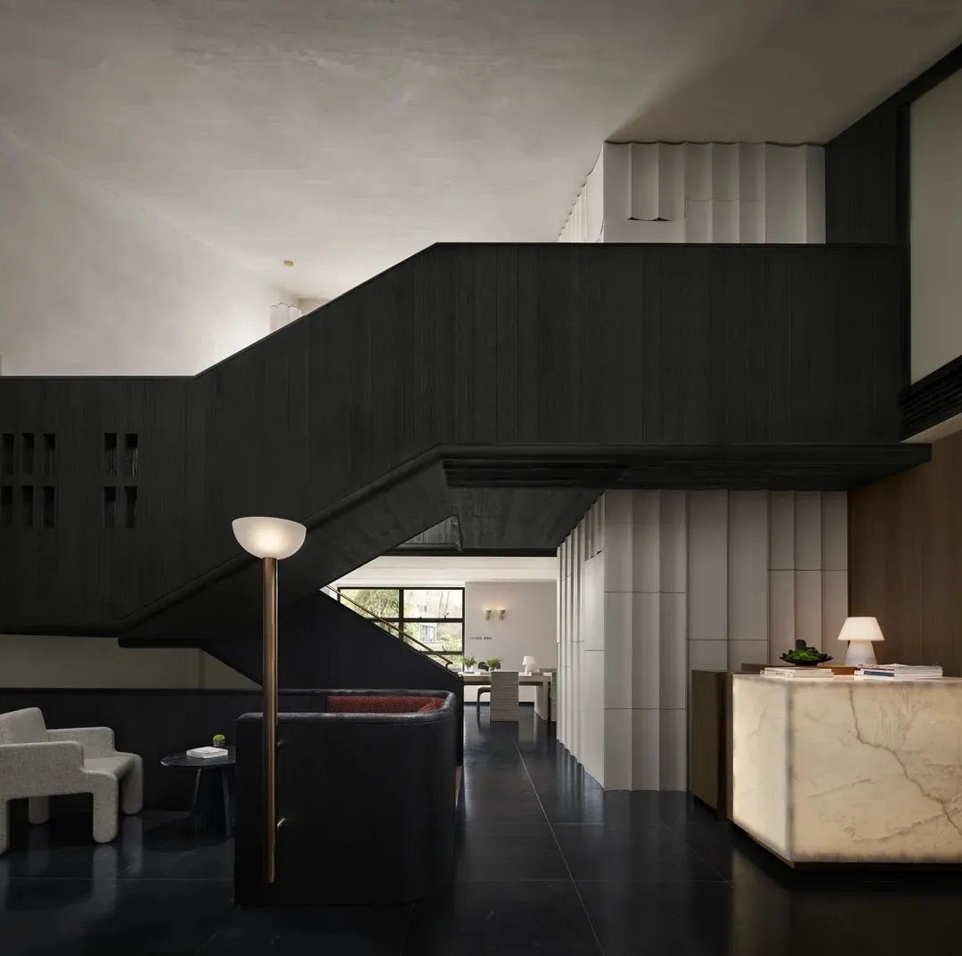 A dimly lit modern living room with a wooden staircase, black railing, and black and white color scheme, featuring two couches and a chair arranged around the staircase