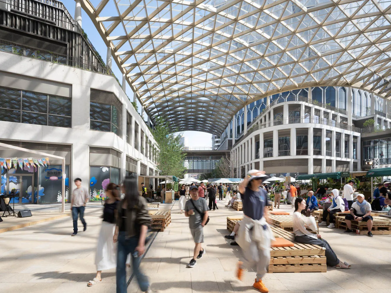 A large, open public space with a glass ceiling, numerous pillars, and a lively atmosphere, filled with people walking, sitting, and engaging in activities
