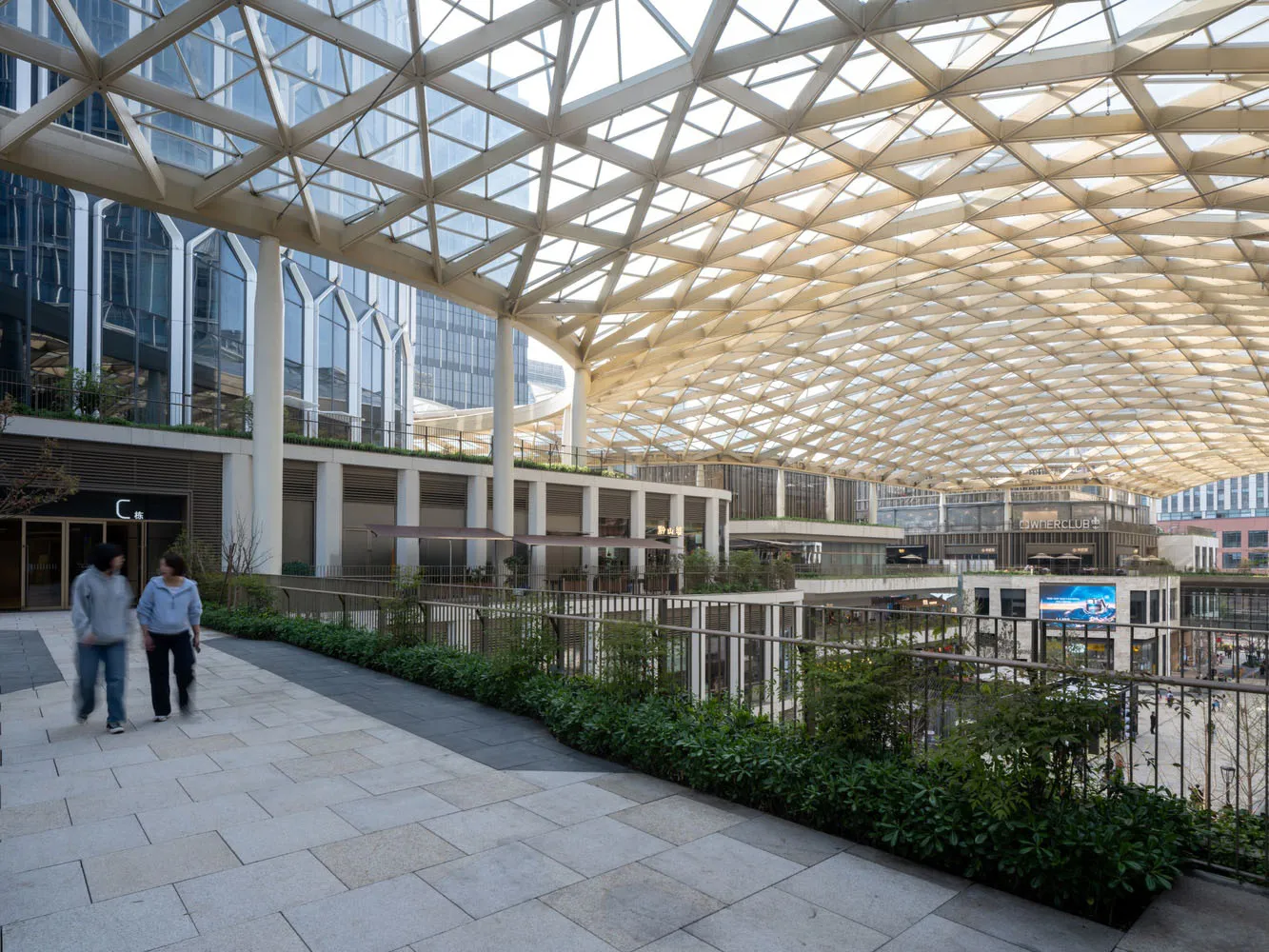 Spacious interior with a mix of modern and traditional architectural styles, featuring a glass ceiling, potted plants, benches, and chairs, with natural light streaming in