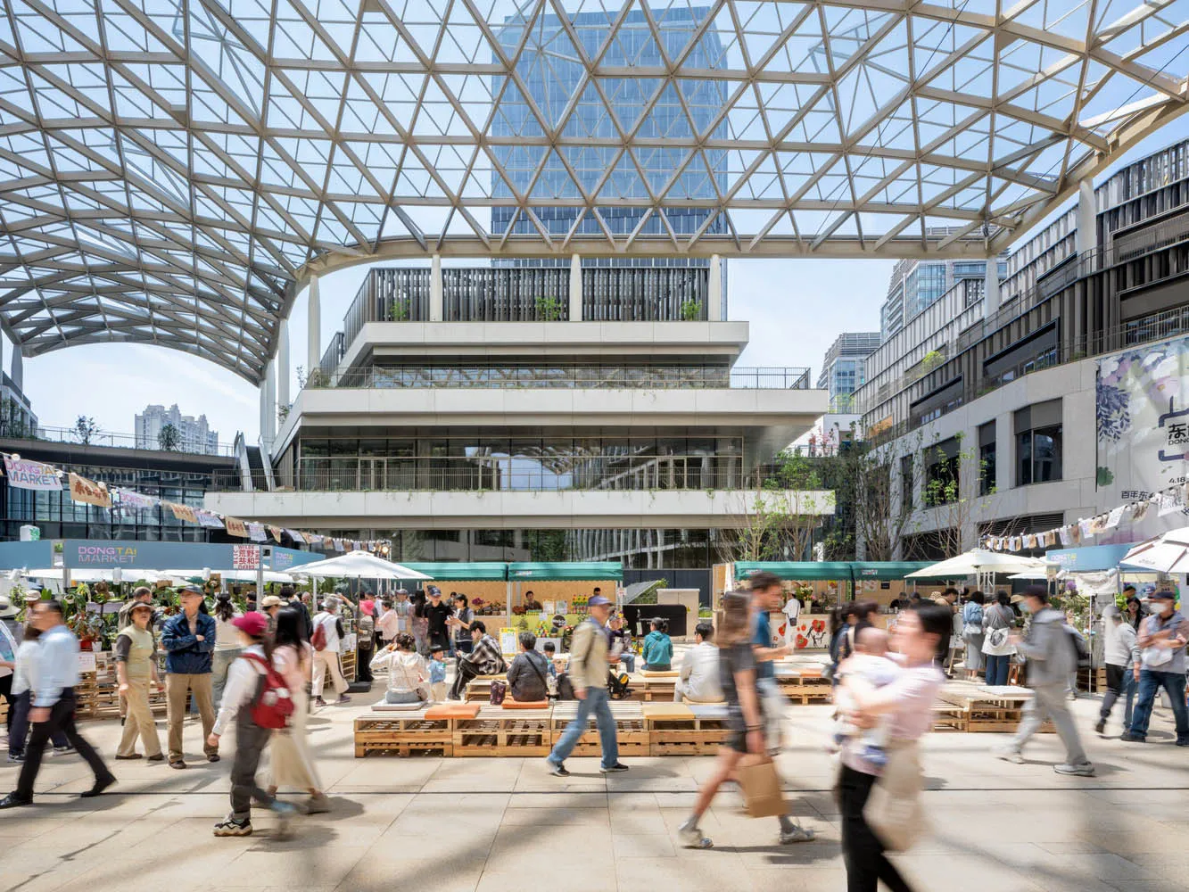 A bustling city street with people walking in various directions, lined with buildings and umbrellas, suggesting outdoor seating or vendors