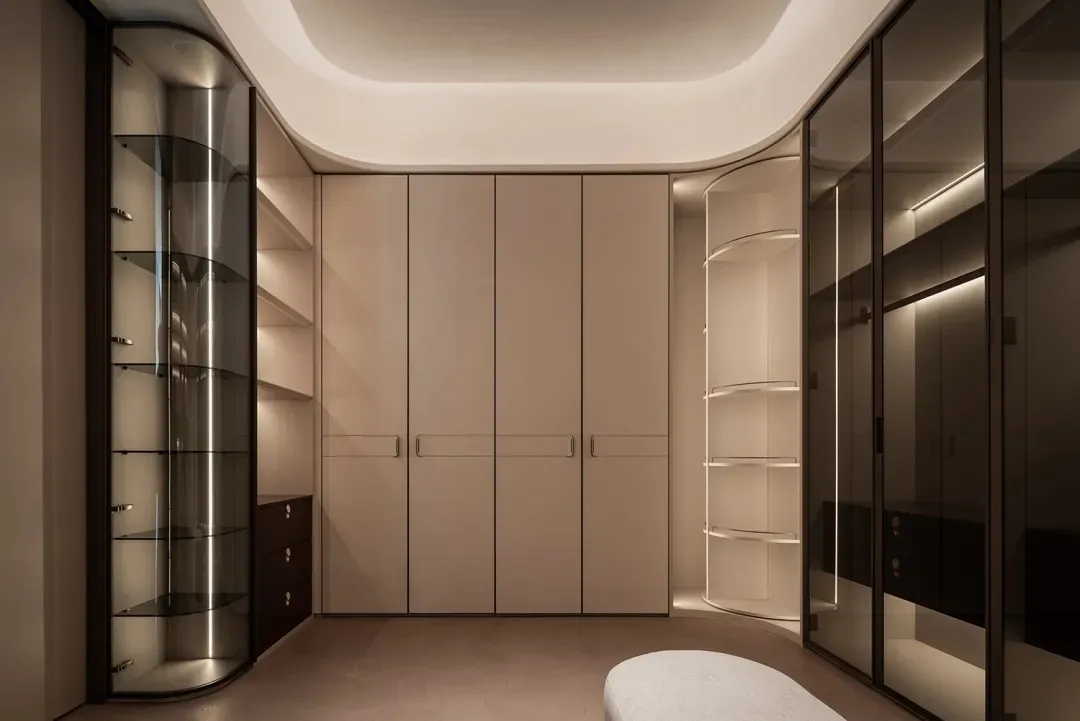 Cozy modern bedroom with a white color scheme, a large closet with a mirrored door, a bookshelf with a glass door, and a bench, set against a dimly lit background