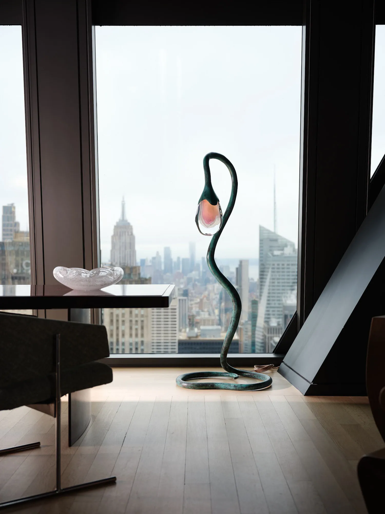 A modern living room with a large window showcasing a cityscape view, featuring a dining table, chair, couch, potted plant, and various decorative items, with natural light creating a bright and inviting space