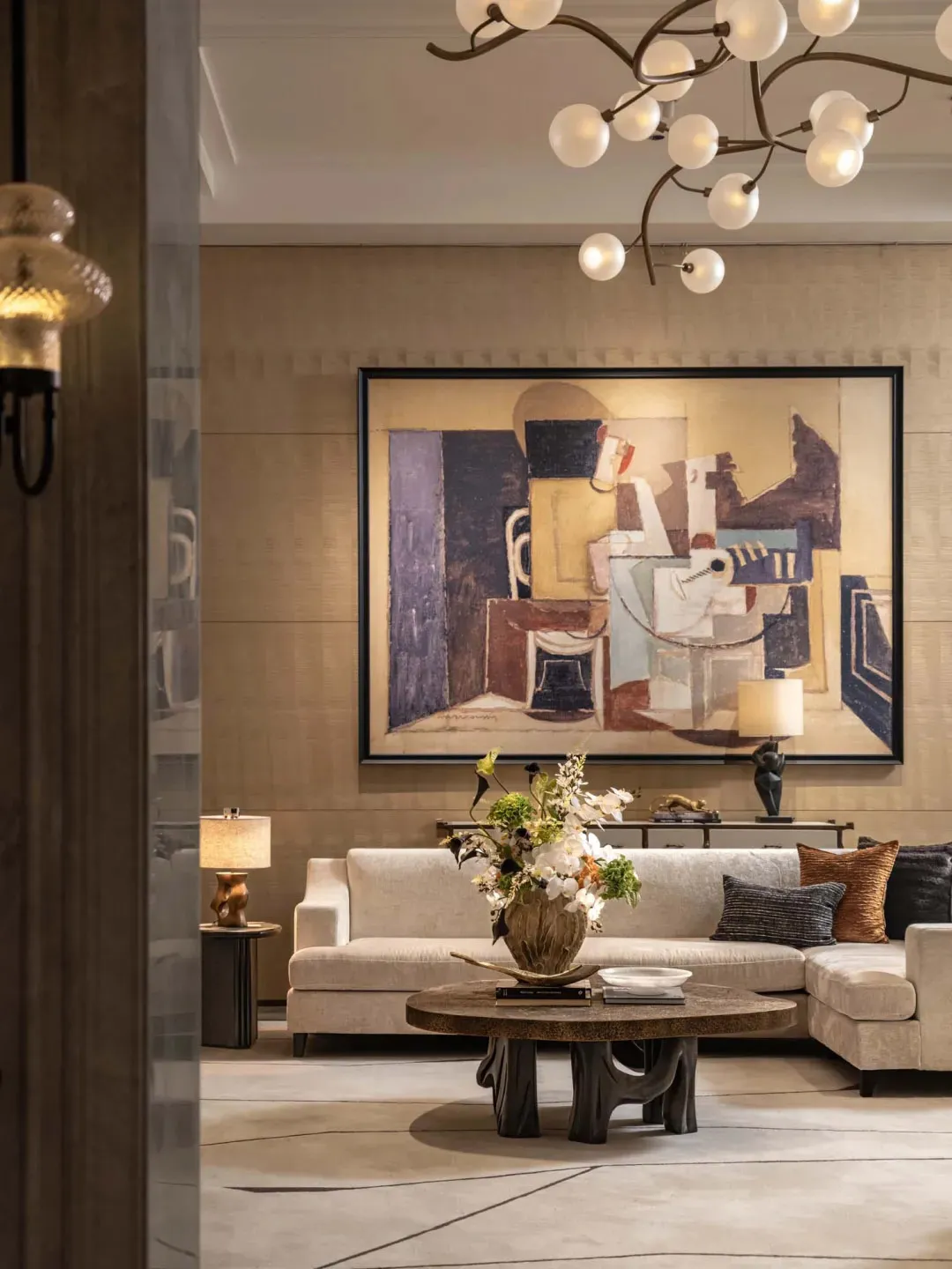 A modern living room with a couch, chair, and dining table, featuring a large painting, wood furniture, and glass accents, with natural and lamp lighting, creating a warm and inviting atmosphere