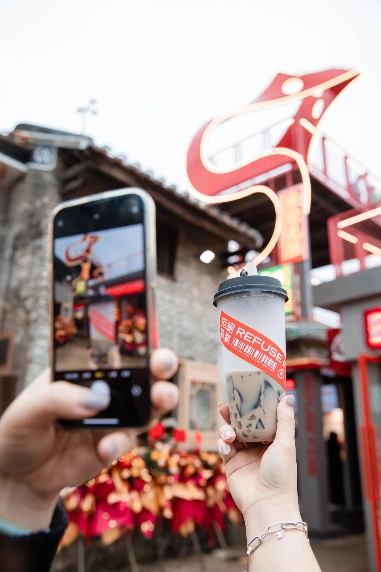A person holding a cup of tea in front of a restaurant, taking a photo with their phone, in a casual outdoor setting with a bench nearby