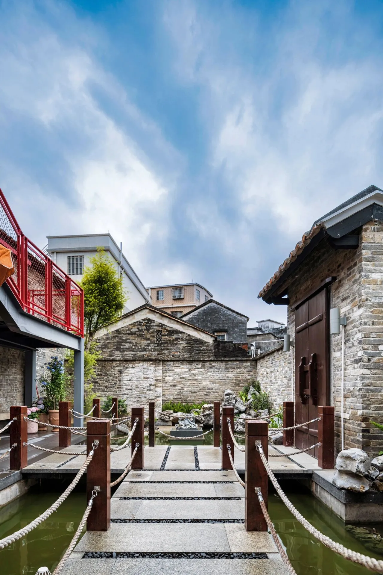 寧靜的庭院，石造建築、磚牆與木橋環繞，盆栽植物與池塘點綴其間，沐浴在自然陽光下