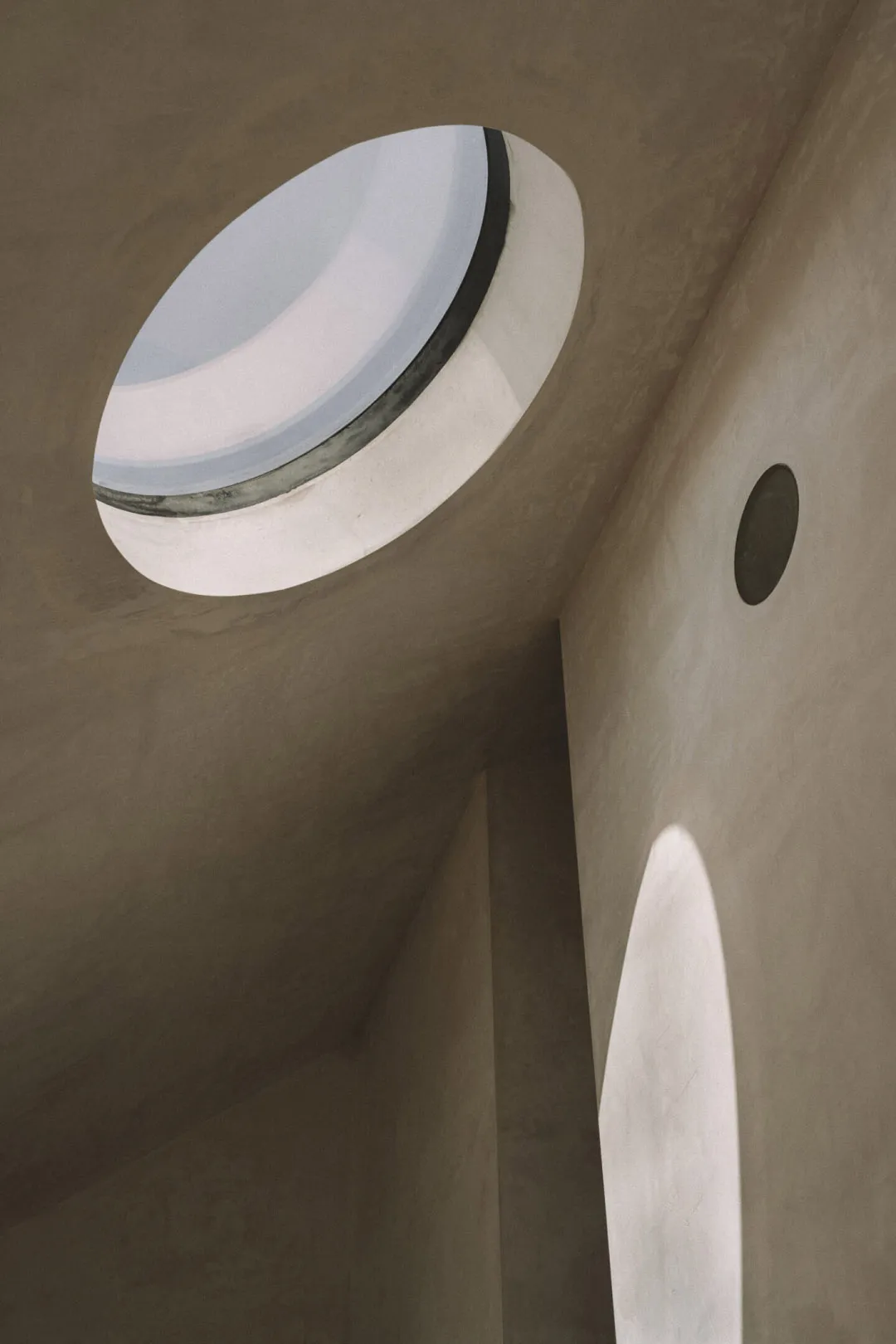 A small, dark room with a round window surrounded by a white frame, set against a concrete-like textured background