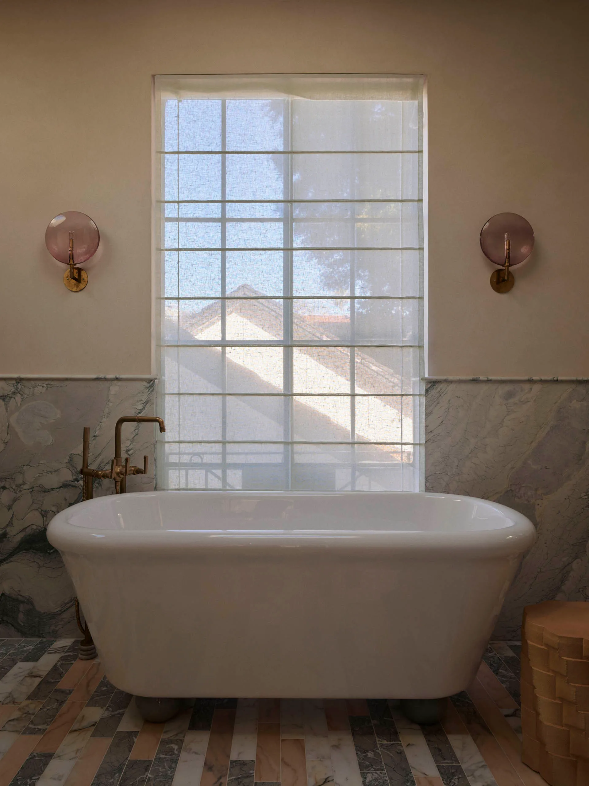 Bathroom with a large white bathtub under a window, surrounded by marble-like surfaces and white curtains, lit by natural sunlight