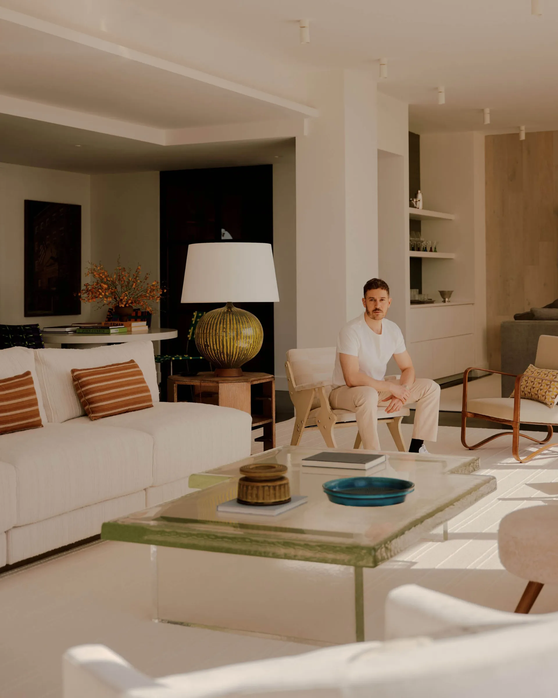 A well-furnished living room with a man sitting on a chair, surrounded by a couch, dining table, and vase, with a comfortable atmosphere