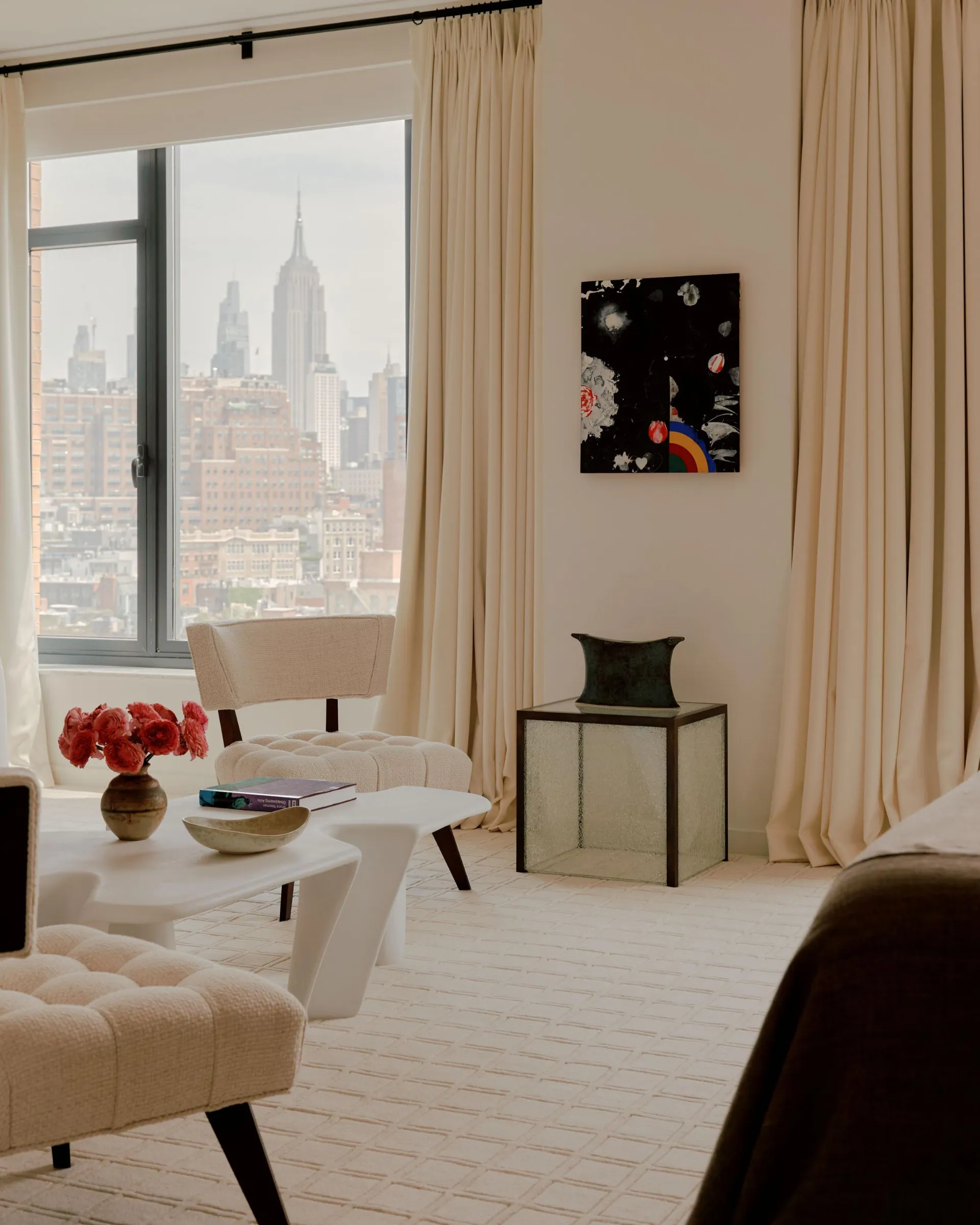 A modern living room with a large window overlooking the city, featuring a couch, chair, and dining table, with a vase and potted plant adding greenery to the space, lit by natural light