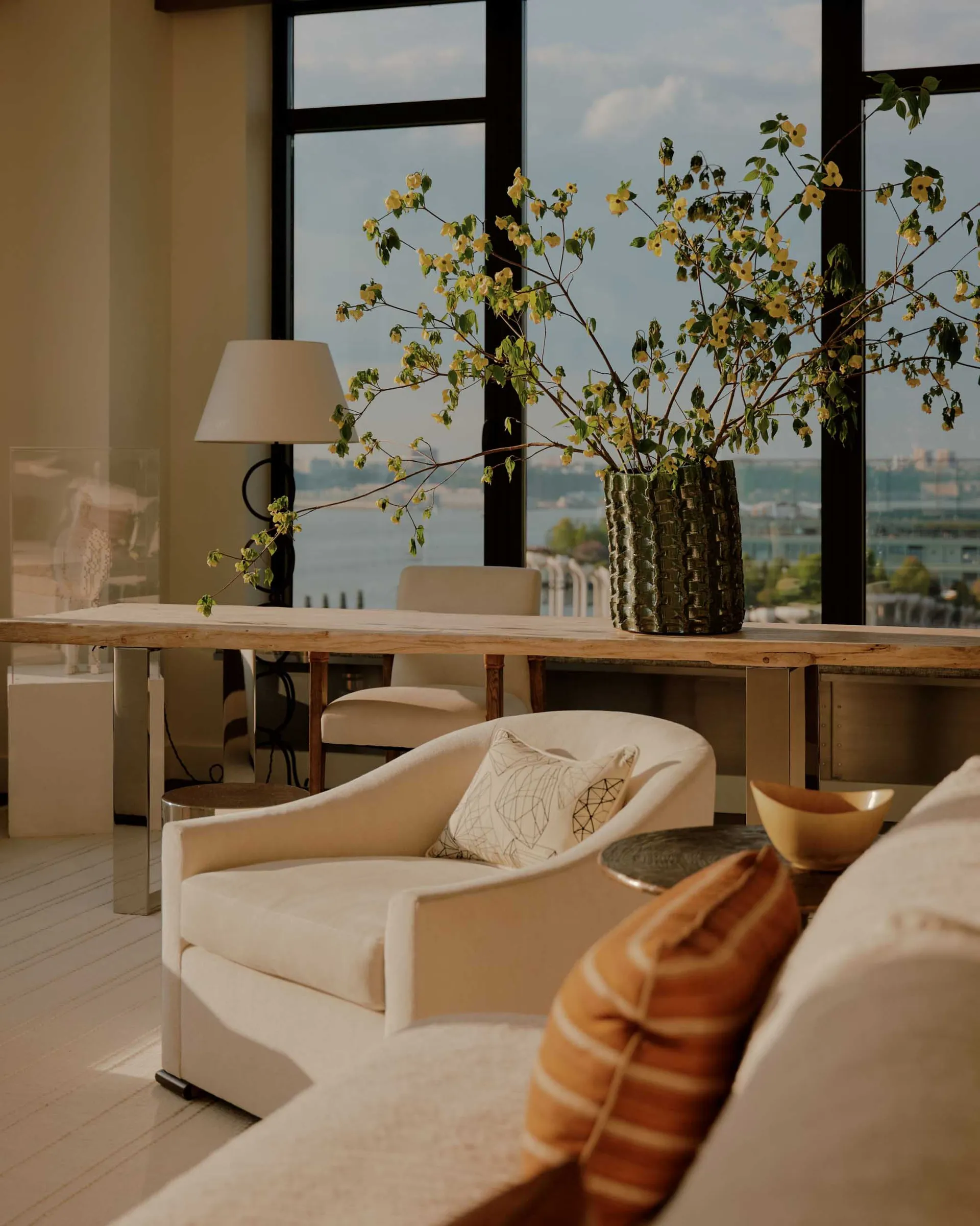 A modern living room with a large window showcasing a city view, featuring a couch, chair, and dining table, with a potted plant adding greenery to the space, and a predominantly white color scheme with wood and glass accents