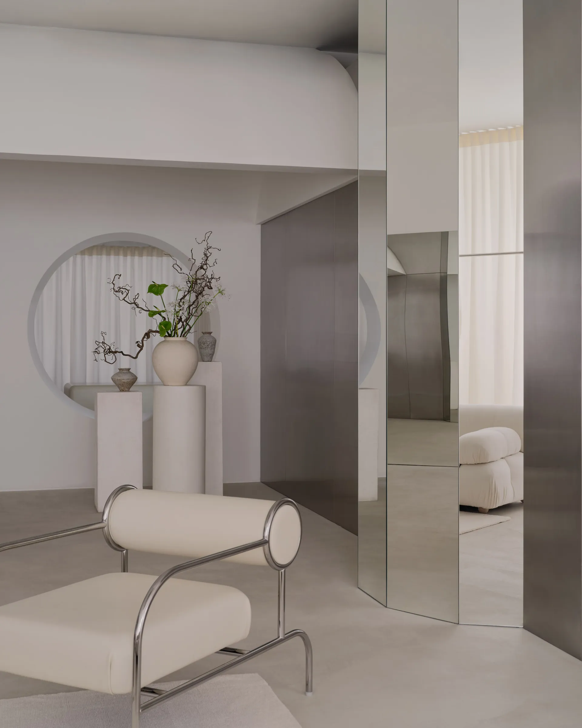 A modern living room with a large mirror, a couch, a chair, and a potted plant, featuring white and grey tones and soft inviting lighting