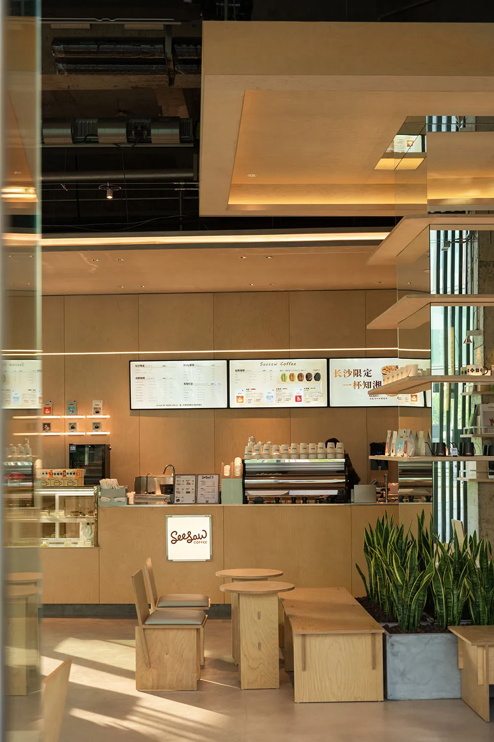 A modern restaurant interior with a dining area and kitchen, featuring wooden elements, a coffee machine, and a refrigerator, with soft and warm lighting