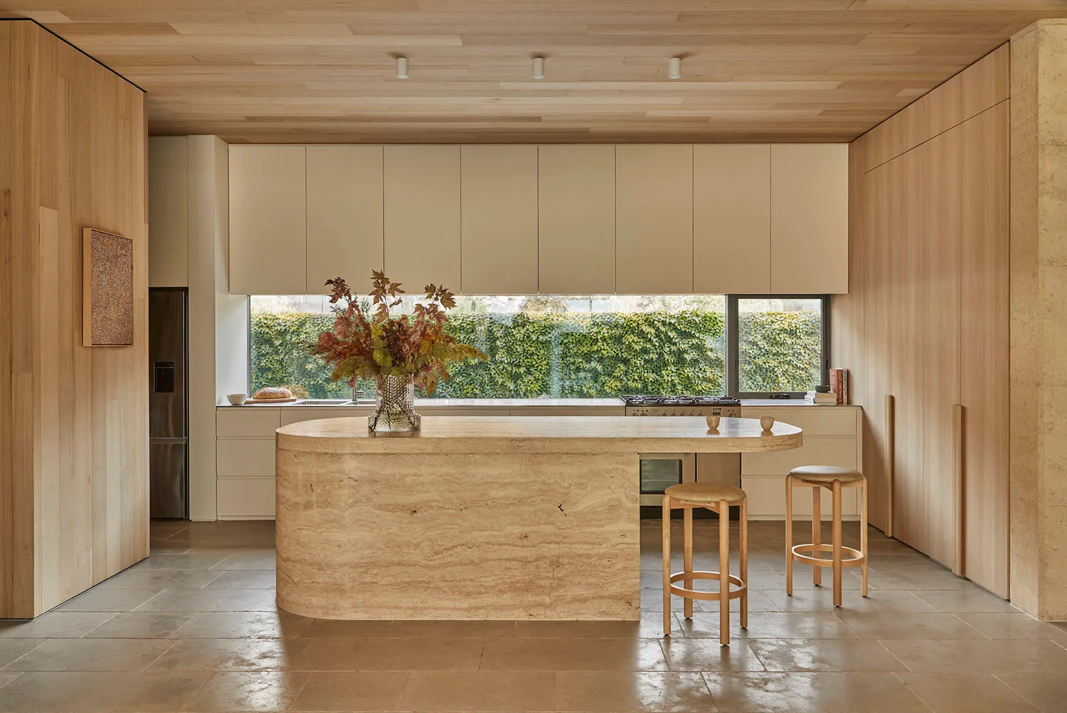 A well-lit kitchen with a marble countertop, wooden cabinets, a sink, refrigerator, and stools, creating a bright and inviting atmosphere