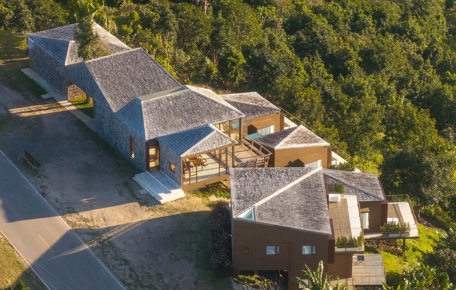 A modern house with a triangular roof, surrounded by trees, featuring a well-lit interior with a dining table and chairs, and a blend of natural materials and indoor-outdoor spaces