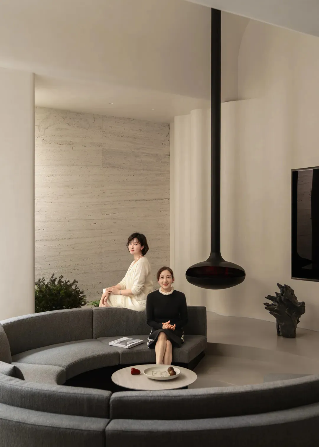 Two women sitting on a couch in a well-lit modern living room with a dining table and potted plant, surrounded by white materials