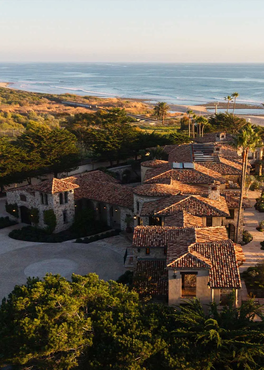 A large Spanish-style house with a red tile roof and various windows, situated on a hillside overlooking the ocean, bathed in natural light