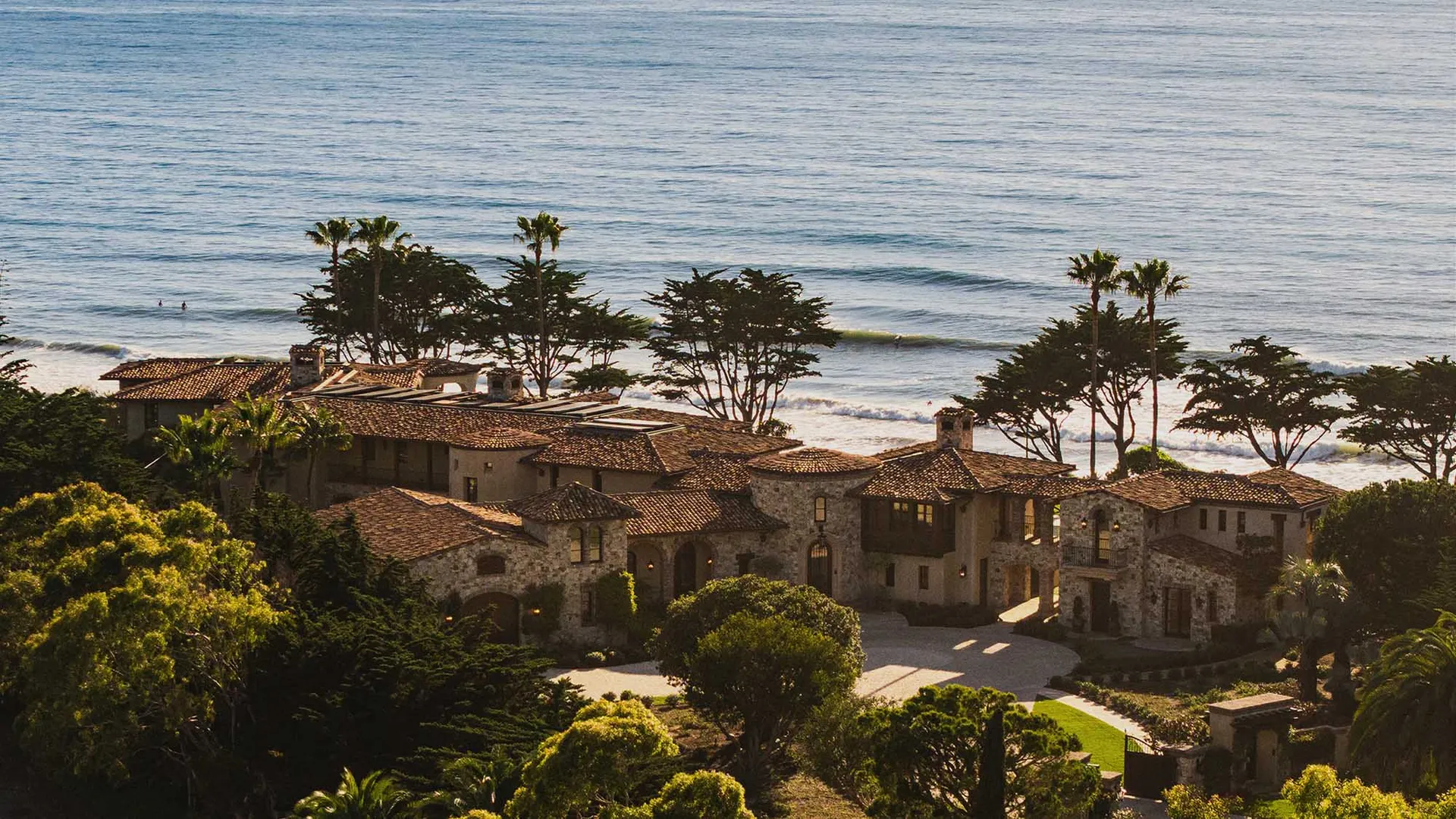 A large Spanish-style house with a red roof and white stucco walls, surrounded by lush greenery and overlooking the ocean