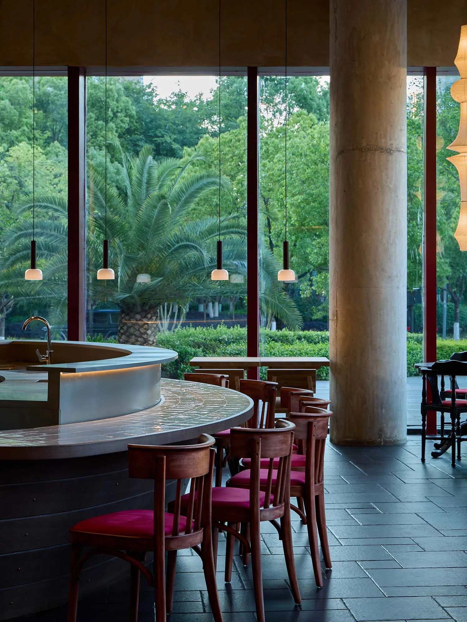 A modern restaurant with a bar, wooden chairs with red cushions, and a large window offering a view of trees outside