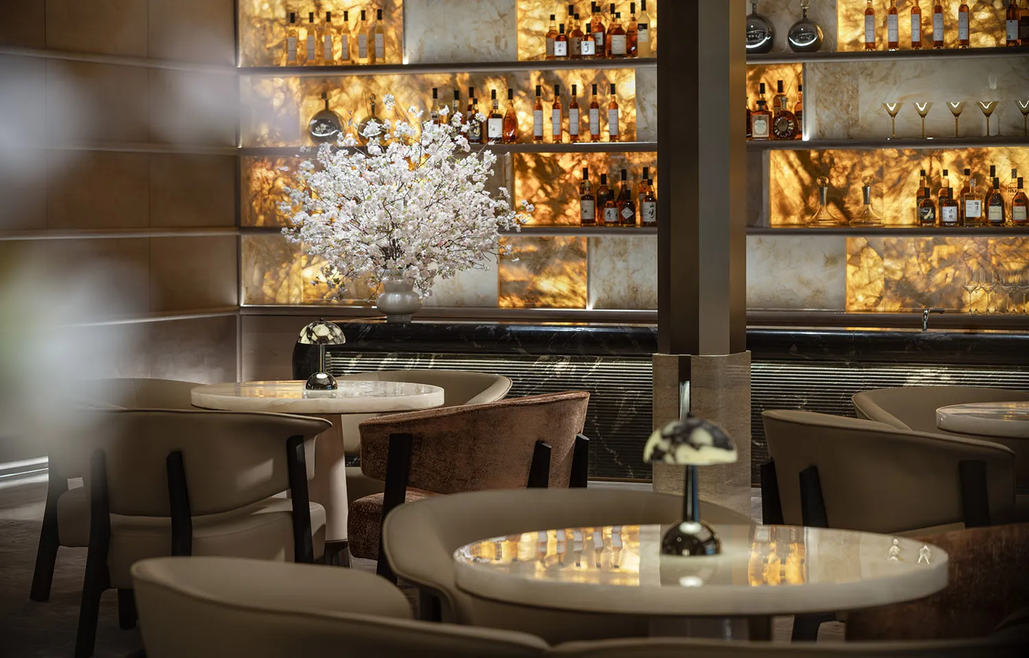 A dimly lit bar with a brown and white color scheme, featuring a counter, bottles on shelves, chairs, and a dining table