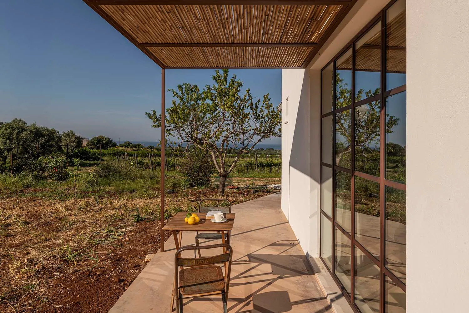 A modern outdoor dining space with a table, chairs, and a shade structure with a woven roof, surrounded by a fence in a residential coastal area