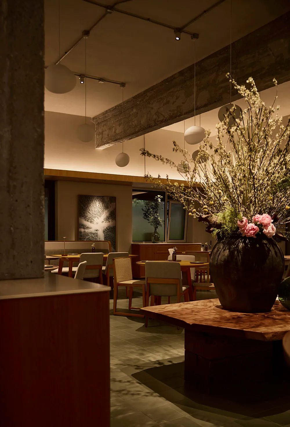 Interior view of a modern wabi-sabi style restaurant featuring wooden tables, upholstered chairs, exposed concrete elements, and a large floral centerpiece in a dark ceramic vase.